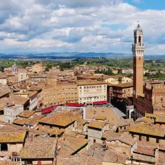 Vista aerea di Siena con la Torre del Mangia che domina la Piazza del Campo, circondata da tetti rossi e colline verdi.