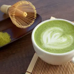 Tazza di matcha latte con schiuma decorata a forma di cuore, accanto a polvere di tè verde e frusta tradizionale.