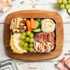 A bento box with cheese, grapes, crackers, nuts, hummus, carrots, and cucumber. Perfect for when are bento boxes eaten cold.