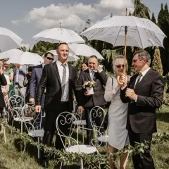 Wesele w kwietniu pod białymi parasolami. Goście w eleganckich strojach czekają na ceremonię na trawie, wśród zieleni i ozdobnych krzeseł.