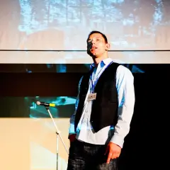 Aktor w kamizelce i koszuli, z identyfikatorem "HAMLET", zastanawia się nad pytaniem: być albo nie być.