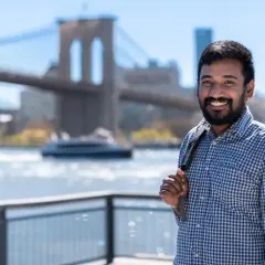 A smiling man with a beard, wearing a blue checkered shirt, stands with the Brooklyn Bridge in the background. He's a student in the Pace University MPA program.
