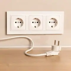 Biały kabel z wtyczką leży na drewnianym blacie, obok gniazdek elektrycznych w ścianie.
