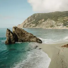 Un rocher émerge des eaux turquoise d'une crique. Une plage de galets s'étend vers un village coloré niché sur une colline verdoyante. Idéal pour savoir où partir en vacances cet été.