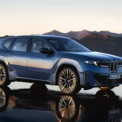 Niebieski BMW Neue Klasse, SUV z futurystycznym designem, stoi na mokrej nawierzchni z odbiciem gór w tle.