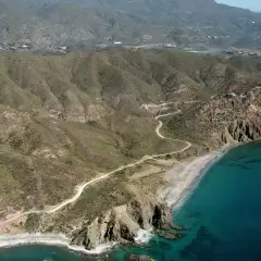 Vista aérea de la playa el lance, con su arena dorada, aguas turquesas y acantilados rocosos. Un camino serpentea por las colinas áridas.