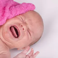Płaczące dziecko z różowym kocykiem na głowie, z otwartymi ustami i zamkniętymi oczami.