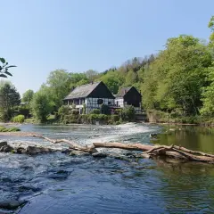 Fachwerkhäuser am Flussufer, wo die **Wipperkotten wandern**. Ein Baumstamm liegt im rauschenden Wasser.