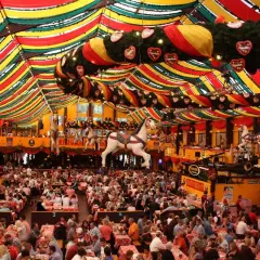 Un tendone colorato ospita la vivace **movida a Monaco di Baviera**. Migliaia di persone siedono ai tavoli, sotto decorazioni a forma di cuore e cavalli da giostra.