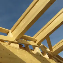 Structure en bois d'une charpente fermette sous un ciel bleu. Les poutres de bois clair forment un motif géométrique complexe.