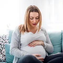 Kobieta w ciąży siedzi na kanapie, trzymając się za brzuch. Jej twarz wyraża dyskomfort, być może to skurcze przepowiadające.