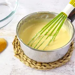 Un batidor verde mezcla una crema amarilla en una olla, mostrando cómo espesar un puré. Cerca hay una cuchara de madera y un recipiente de vidrio.