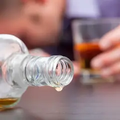 Ostatnia kropla alkoholu metylowego kapie z butelki. W tle niewyraźna postać trzyma szklankę z bursztynowym płynem.