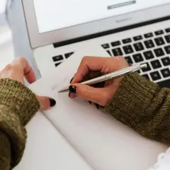 Dłonie piszące w notatniku obok laptopa.