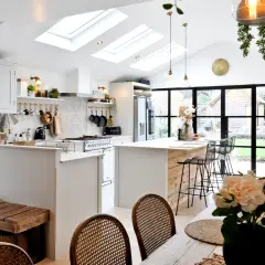 Jasna kuchnia w stylu modern farmhouse z wyspą, białymi meblami i drewnianymi akcentami. Dachowe okna wpuszczają mnóstwo światła.