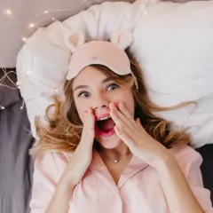 En tjej med sovmask och rosa pyjamas ligger i sängen, redo för att sova med sminket kvar. Hon ser överraskad ut.