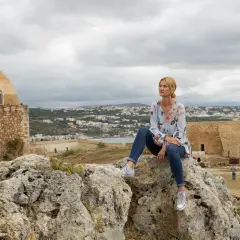 Kobieta siedzi na skale, podziwiając widok miasta. Kreta pogoda październik, chmury na niebie.