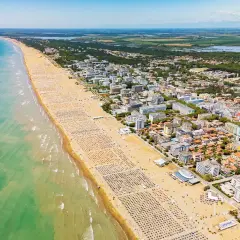 Słoneczny dzień nad morzem. Tłumy plażowiczów na szerokiej, piaszczystej plaży, a za nią miasteczko z hotelami i zielenią.