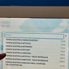 Palec wskazuje opcję "PIERWSZA REJESTRACJA SAMOCHODU" na ekranie terminala, gdzie można uzyskać upoważnienie wydział komunikacji.