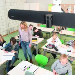 Lehrerin betreut Schüler im Klassenzimmer, die an ihren Tischen arbeiten. Ein modernes Setup für hybriden Unterricht.
