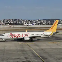 Samolot linii lotniczych Pegasus z napisem "flypgs.com" na kadłubie na płycie lotniska, w tle miasto.