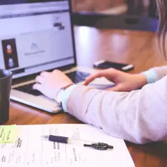 Kobieta pracuje przy laptopie. Na biurku kubek, notatki i długopis. Oświetlenie zapewnia odpowiednie ile luxów na stanowisku pracy.