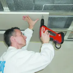 Mężczyzna w białej kurtce montuje płyty gk do stelaża sufitowego za pomocą wkrętarki.