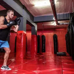A boxer trains in a gym with many boxing bags. The red floor reflects the lights, and the boxing bag numbers are not visible.