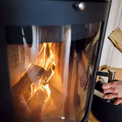 Eine Hand legt Holzscheite in einen brennenden Ofen. Die Flammen lodern im Inneren des Ofens.