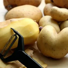 Ziemniaki, bogate w węglowodany, są obierane za pomocą obieraczki. Widać skórki i kilka ziemniaków.