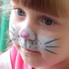 Ein kleines Mädchen mit blauem Auge und Kinderschminken als Tier. Die Nase ist rosa, die Wangen mit Punkten und Schnurrhaaren bemalt.
