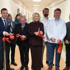 Grupa osób w garniturach i kitlach przecina czerwone wstęgi w szpitalnym korytarzu.