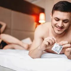 Mężczyzna w łóżku przygotowuje się do intymnego spotkania, trzymając w dłoni prezerwatywę. Kobieta leży w tle. Rozmiary prezerwatyw są kluczowe dla komfortu.
