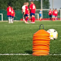 Stos pomarańczowych pachołków i piłka nożna na zielonej murawie, w tle dzieci w strojach piłkarskich.