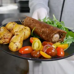 Une galette saucisse roulée, servie avec des pommes de terre rôties et des tomates cerises colorées, sur une assiette noire.