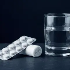 Confezione di farmaci per mal di denti, rotolo di garza e bicchiere d'acqua su sfondo scuro.