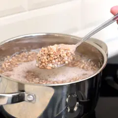 Eine Hand hebt einen Löffel mit dampfendem Buchweizen aus einem Topf, in dem Buchweizen gekocht wird.