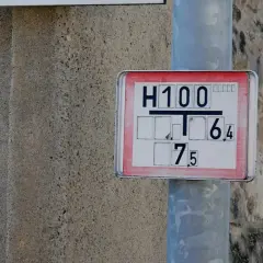 Nahaufnahme eines Hydrantenschilds mit der Aufschrift "H100 T 6,4 7,5". Das Schild ist an einem Metallpfosten befestigt, daneben eine raue Betonwand.