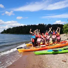 Grupa przyjaciół w kamizelkach ratunkowych pozuje przy kajakach na plaży. W tle widać wodę i zalesione wyspy archipelagu sztokholmskiego.