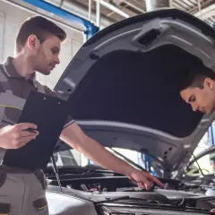 Dwóch mechaników sprawdza silnik samochodu. Jeden z nich trzyma notatnik, drugi wskazuje na element pod maską.