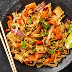 Talerz pełen makaronu ryżowego z warzywami i tofu, polany aromatycznym sosem pad thai. Obok pałeczki i ćwiartka limonki.