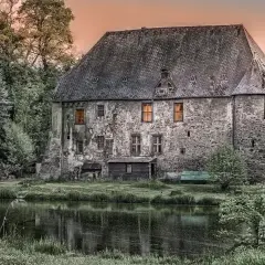 Kamienny dworek szlachecki z oknami, otoczony drzewami, nad stawem.