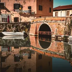 Un ponte in mattoni rossi attraversa un canale nel quartiere Venezia di Livorno. Barche ormeggiate riflettono sulle acque calme, mentre panni stesi aggiungono un tocco di vita quotidiana.