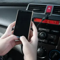 Dłonie trzymają telefon, który jest podłączony kablem do radia samochodowego. Prosty sposób, jak połączyć telefon z samochodem.