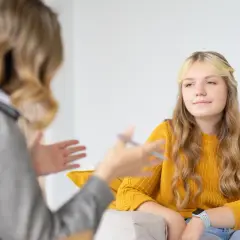 Nastolatka w żółtym swetrze podczas terapii. Rozmowa z terapeutką, która gestykuluje.