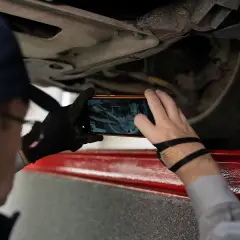 Mechanik sprawdza podwozie samochodu, używając telefonu do zrobienia zdjęcia. Szuka, gdzie jest kod silnika, aby zidentyfikować usterkę.