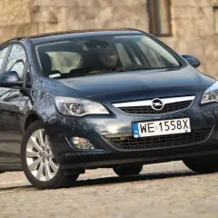 Ciemny Opel Astra J z włączonymi światłami do jazdy dziennej, zaparkowany na kamienistej drodze.