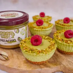 Sześć mini serniczków z kremem pistacjowym, posypanych pistacjami i ozdobionych maliną. Obok słoik kremu pistacjowego.