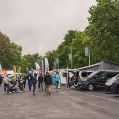 Ludzie zwiedzają wystawę kamperów i przyczep. W tle drzewa i flagi. To zlot caravaningowy.