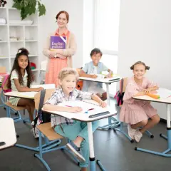 Uśmiechnięta nauczycielka z książką "English grammar" i dzieci przy ławkach. Widać, że to świetny moment, kiedy najlepiej się uczyć.